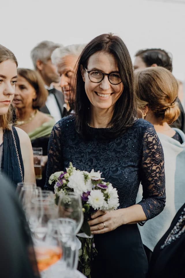 Beatrix Diedenhofen lächelt mit einem Blumenstrauß in der Hand inmitten von Gästen bei einem Fest