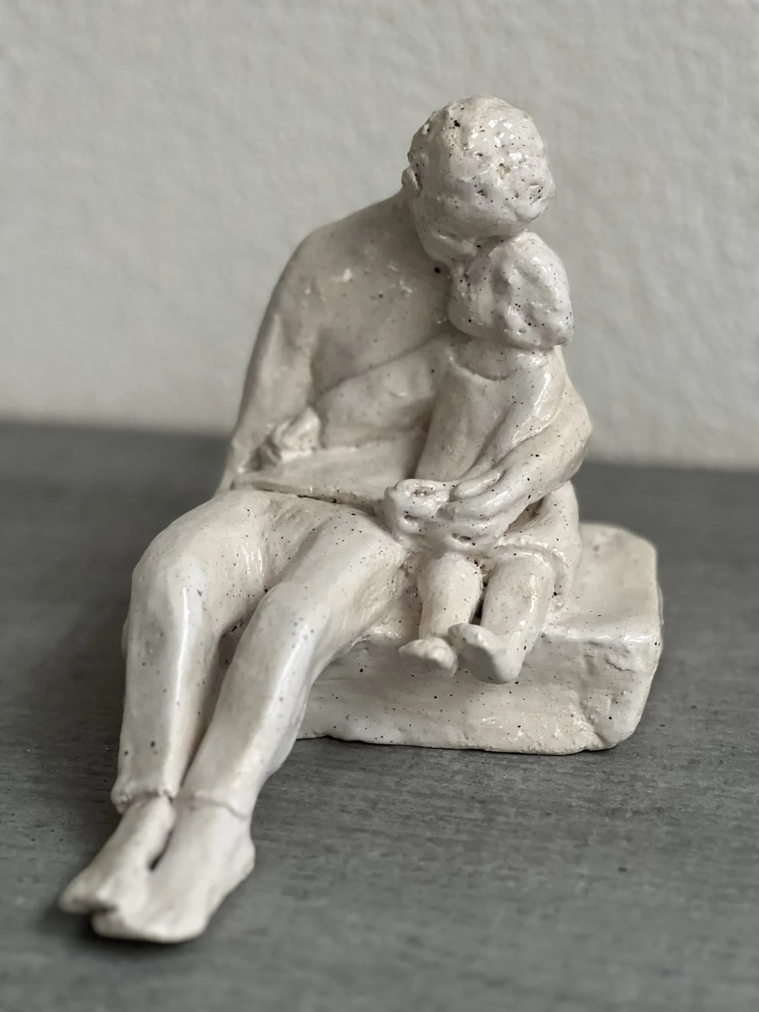 Weiß glasierte Keramikskulptur eines sitzenden Elternteils mit einem Kind auf dem Schoß.