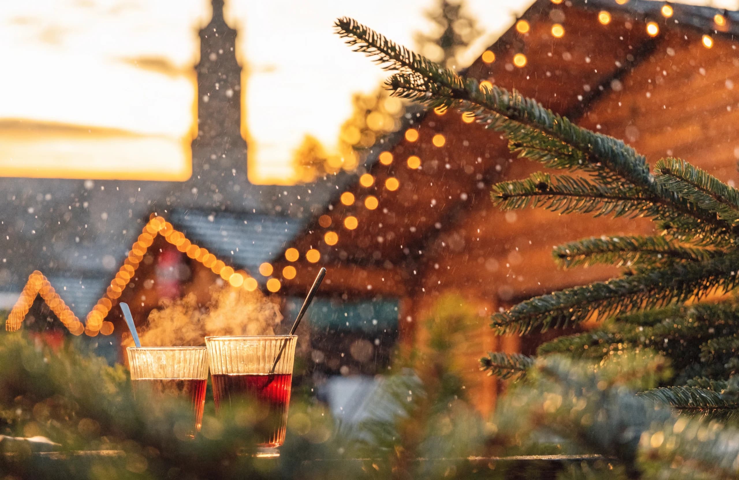 Winterlicher Weihnachtsmarkt in der Dämmerung mit Lichterketten an Holzständen, Tannenzweigen und zwei dampfenden Glühwein-Tassen im Vordergrund.